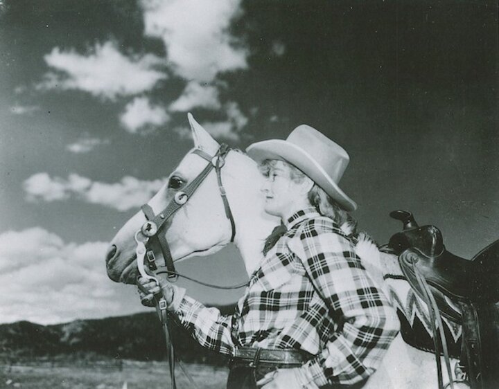Black and white photo of Mari Sandoz. She wears a plaid shirt and cowboy hat. She stands next to a horse.xt to a horse.
